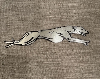 May include: A silver-toned metal decal featuring a stylized silhouette of a greyhound dog in a full running stride. The design includes black engraved-style lines to define the musculature, facial features, and tail of the dog in motion.