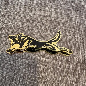 May include: A metallic gold and black sticker featuring a silhouette of a running dog with its mouth open in a growling expression. The design shows the dog in a dynamic, horizontal pose with detailed fur markings and a pointed tail.