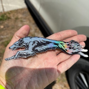 Könnte beinhalten: Ein glitzernder, stilisierter Wolfsaufkleber in Laufpose. Der Wolf ist hauptsächlich silber und schwarz, mit schillernden Regenbogenakzenten entlang seines Schwanzes. Der Aufkleber wird in der offenen Hand einer Person gehalten, mit verschwommenem Hintergrund.