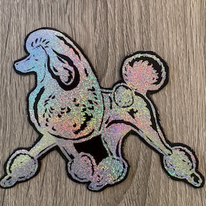 May include: A holographic sticker shaped like a poodle in a classic continental clip. The design features a shimmering, multi-coloured glitter finish with bold black outlines defining the dog's curly fur, pom-poms on the legs, and tail.