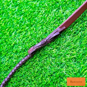 May include: A brown leather riding crop with a braided handle and decorative accents. The crop is set against a backdrop of bright green artificial grass, with the word "Bantoro" in a small orange rectangle.
