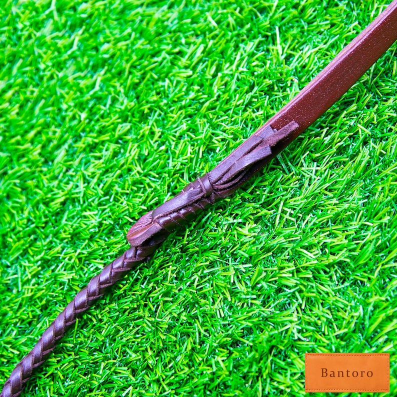 May include: A brown leather riding crop with a braided handle and decorative accents. The crop is set against a backdrop of bright green artificial grass, with the word "Bantoro" in a small orange rectangle.