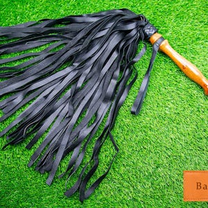 Puede incluir: Un flagelador de cuero negro con un mango de madera. Las hebras del flagelador son largas y finas, y el mango es de color marrón claro. El flagelador está sobre una superficie verde. La palabra "Bantoro" está en una etiqueta marrón.