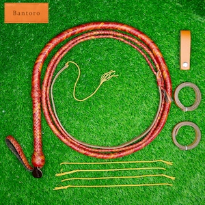 May include: A braided leather whip in shades of red and orange, with a matching handle and accessories. The whip is coiled on a green surface, with additional leather pieces and a tag that reads "Bantoro".