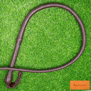 May include: A brown leather whip with a braided handle and loop, resting on a green surface. The whip is made of woven leather and has a knotted end. The word "Bantoro" is printed on a small orange rectangle.