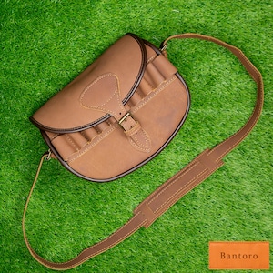 May include: A brown leather shoulder bag with a curved design, featuring a flap closure secured by a brass buckle. The bag has a long, adjustable strap and several loops on the front. The bag is set against a green background. The brand name "Bantoro" is visible.
