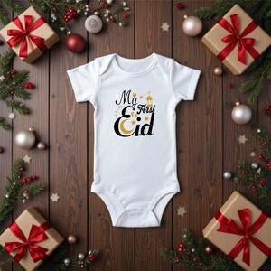 Pu&ograve; includere: Body bianco per neonato con testo nero che recita "My First Eid" con un grafico di luna crescente dorata, stelle e lanterna.