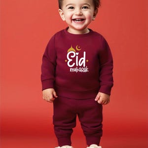 Pu&ograve; includere: Un bambino piccolo che indossa una felpa e pantaloni della tuta abbinati di colore bordeaux con il testo "Eid Mubarak" e un design di mezzaluna e moschea dorati. Il bambino sorride e indossa scarpe da ginnastica bianche.
