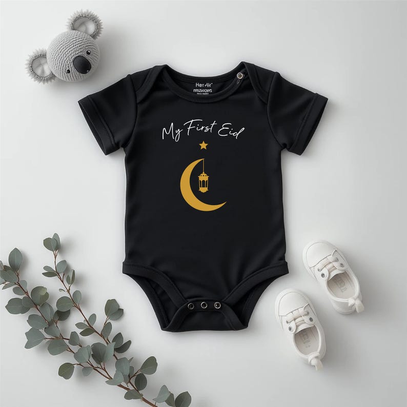 Pu&ograve; includere: Body nero per neonato con maniche corte e testo bianco che dice "My First Eid" con un'immagine di una mezzaluna dorata e una lanterna.