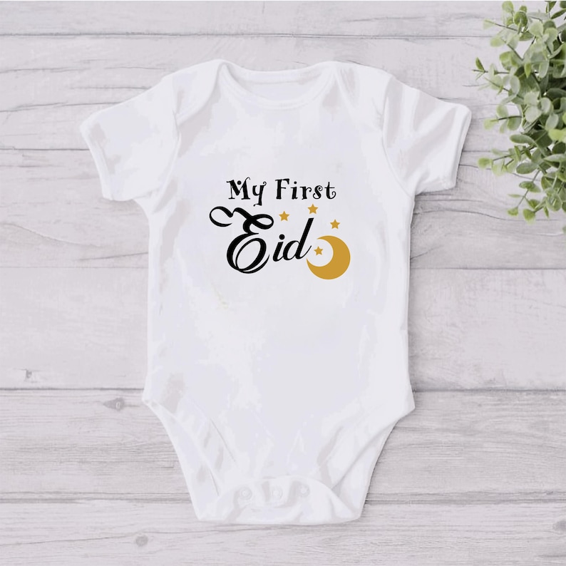 Pu&ograve; includere: Body bianco per neonato con testo nero che recita "My First Eid" con una mezzaluna dorata e stelle.