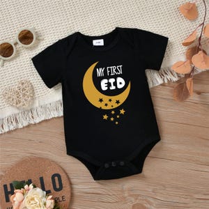 Pu&ograve; includere: Body nero a maniche corte con un motivo a mezzaluna dorata e stelle. Il testo "My First Eid" &egrave; stampato sulla luna.