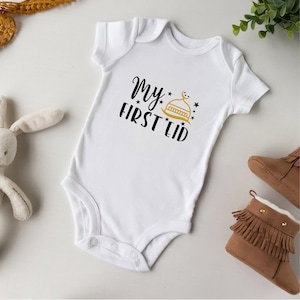 Pu&ograve; includere: Body bianco per neonato con testo nero che recita "My First Eid" e un'illustrazione dorata di una moschea con una mezzaluna e stelle.