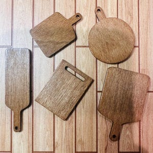 May include: Five wooden cutting boards in various shapes, including rectangular, square, and round, with handles. The boards are made of light brown wood and arranged on a wooden surface.