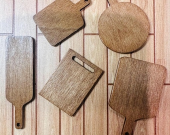 Puede incluir: Cinco tablas de cortar de madera de varias formas, incluyendo rectangulares, cuadradas y redondas, con asas. Las tablas son de madera marrón claro y están dispuestas sobre una superficie de madera.