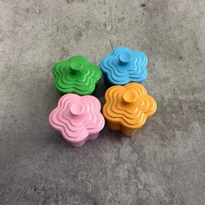 Op de afbeelding: Vier bloemvormige knoppen in groen, blauw, roze en oranje. Elke knop heeft een centrale, cirkelvormige handgreep. De knoppen zijn gerangschikt op een grijze ondergrond en bieden een levendig accent voor meubels of decoratie.