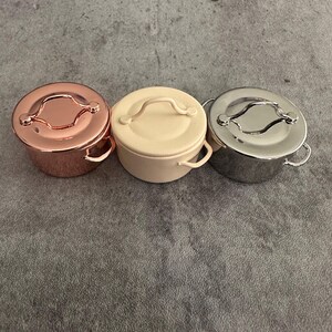 May include: Three small, round metal pots with lids. The pots are in rose gold, cream, and silver. Each pot has a handle on the lid and two small handles on the sides. The pots are sitting on a gray surface.