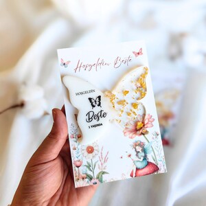 Pode incluir: Um cartão branco com um design de borboleta e detalhes dourados. O cartão tem o texto "Hosgeldin Beste" e "1 Yasinda" impresso nele. O cartão é decorado com flores e uma fada.