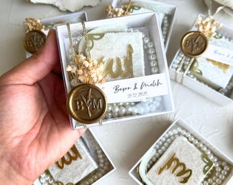 Puede incluir: Cajas de regalo blancas con tapas transparentes que contienen cuadrados de tela blanca con letras doradas, collares de perlas blancas y sellos de cera dorados con las iniciales "B&M". Las cajas tienen una etiqueta con el texto "Basya & Melich 25.12.2023".