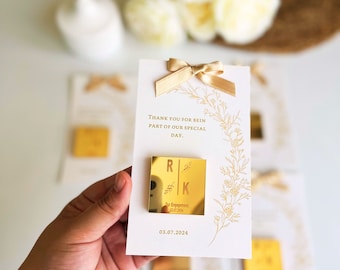May include: Gold mirror favor with the initials "R" and "K" and the text "Our Engagement 03.07.2024" attached to a white card with gold floral accents and the text "Thank you for being part of our special day."