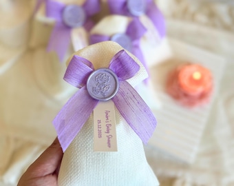 May include: Small, off-white fabric bags tied with lavender ribbons and sealed with a lavender wax stamp featuring a rose design. Each bag has a small tag with text. The bags are likely party favors or gifts.
