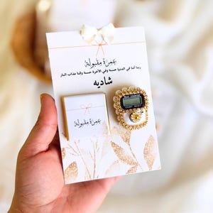 May include: A white card with gold glitter accents and a gold bow. The card has Arabic text that reads "Umrah Mabrour" and "Shadiya". The card is next to a white and gold digital prayer counter with a black display.