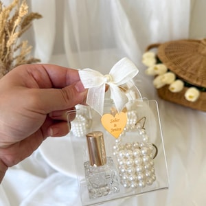 Könnte beinhalten: Ein transparentes Geschenkset mit einer kleinen Parfümflasche mit goldenem Verschluss und einem perlenbesetzten Schlüsselanhänger. Das Set befindet sich in einer transparenten Plastiktüte, die mit einer weißen Schleife und einem herzförmigen goldenen Anhänger mit den Namen "Saber & Jenine" gebunden ist.