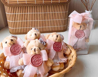 May include: Four small teddy bears wearing pink dresses with a pink ribbon and a wax seal with the word "Little" on it. The bears are in a wicker basket.