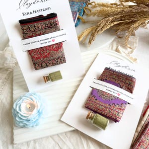 May include: Two small, patterned fabric pouches with black zippers are displayed on white cards. Each card has text that reads "Olaydanın KINA HATIRASI" and "THANK YOU FOR ACCOMPANYING US ON OUR HAPPY DAY". A small glass bottle is also present.