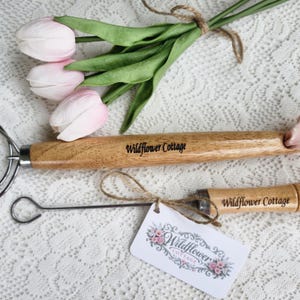 May include: A stainless steel dough whisk and a smaller metal hook tool, both featuring light brown wooden handles engraved with the text Wildflower Cottage. The larger whisk has a pink fabric ribbon tied to the end, and the smaller tool has a white tag attached with the text Wildflower Cottage and a floral graphic.