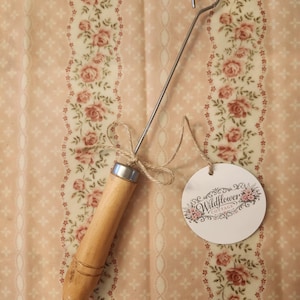 May include: A wooden handled tool with a metal hook and a decorative tag. The tool has a light brown wooden handle with a silver metal band. The tag is white with the words "Wildflower Cottage" and floral accents.