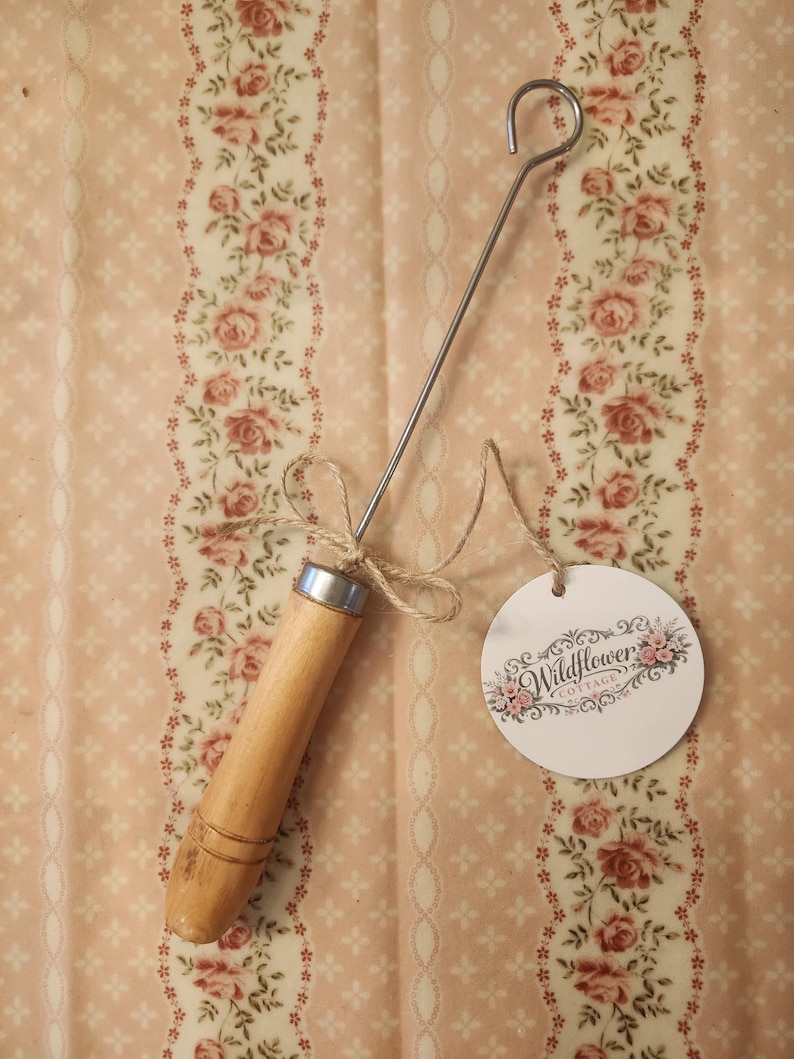 May include: A metal hook tool with a light-colored wooden handle and a silver-toned metal ferrule. A piece of twine is tied around the neck of the handle, attaching a round white paper tag printed with the text Wildflower Cottage and floral art.