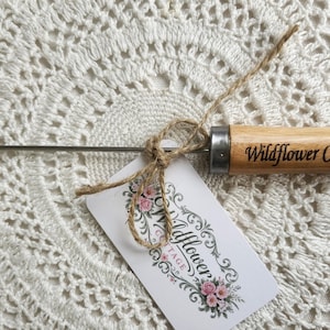 May include: A metal tool with a circular loop at one end and a light brown wooden handle. The handle is engraved with the text Wildflower Cottage. A small white tag with floral graphics and the same text is tied to the tool with twine.
