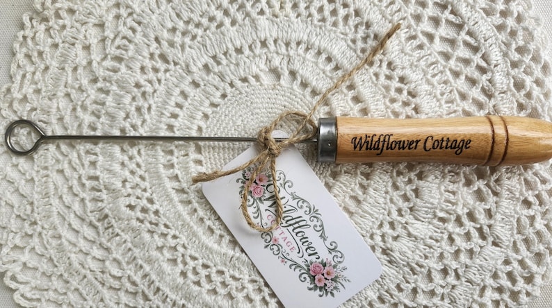 May include: A metal tool with a circular loop at one end and a light brown wooden handle. The handle is engraved with the text Wildflower Cottage. A small white tag with floral graphics and the same text is tied to the tool with twine.