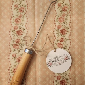 May include: A metal hook tool with a cylindrical wooden handle and a silver-colored metal ferrule. A piece of jute twine is tied around the handle, attaching a round white paper tag printed with the text Wildflower Cottage and floral graphics.
