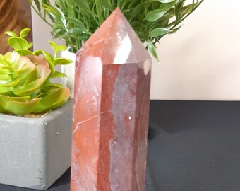 May include: A red and white crystal point with a faceted shape. The crystal is sitting on a black surface.