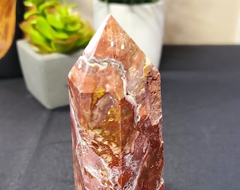 May include: A red, white, and brown crystal point with a natural, rough texture. The crystal is shaped like a pyramid and has a pointed top.