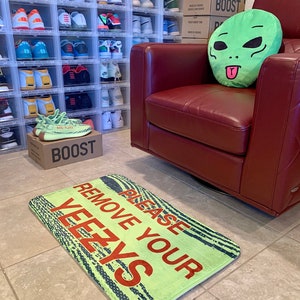 May include: A green alien-shaped pillow sits on a red leather armchair. A green doormat with the text "Please Remove Your Yeezys" is on the floor. The doormat has a black and white striped pattern. There are boxes stacked on the wall behind the chair that say "Boost" on them.