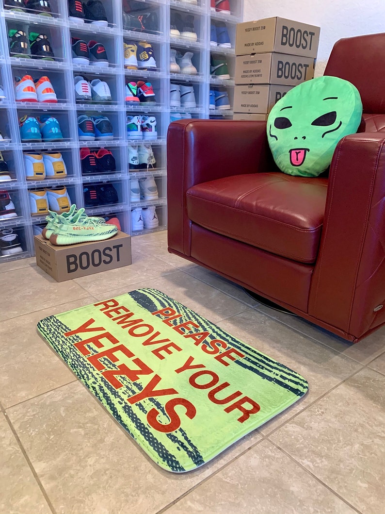 May include: A green alien-shaped pillow sits on a red leather armchair. A green doormat with the text "Please Remove Your Yeezys" is on the floor. The doormat has a black and white striped pattern. There are boxes stacked on the wall behind the chair that say "Boost" on them.