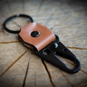 Peut inclure: Un porte-clés en cuir noir avec un clip en métal et une boucle en cuir marron. Le porte-clés est sur une surface en bois.