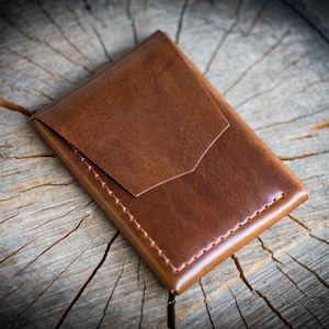 Peut inclure: Un porte-cartes en cuir marron avec une finition lisse, une fermeture à rabat pointu et des coutures visibles sur les bords. Cet accessoire rectangulaire est conçu avec une poche unique et présente une couleur brun clair uniforme sur l'ensemble.