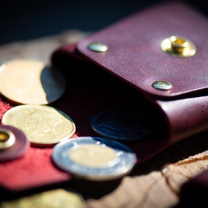 K&ouml;nnte beinhalten: Eine bordeauxrote Lederm&uuml;nzb&ouml;rse mit einer Klappe und einem Druckknopfverschluss. Die B&ouml;rse ist offen und mehrere Gold- und Silberm&uuml;nzen fallen heraus.