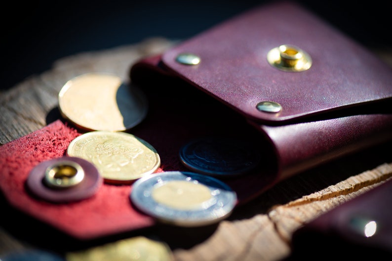 K&ouml;nnte beinhalten: Eine bordeauxrote Lederm&uuml;nzb&ouml;rse mit einer Klappe und einem Druckknopfverschluss. Die B&ouml;rse ist offen und mehrere Gold- und Silberm&uuml;nzen fallen heraus.