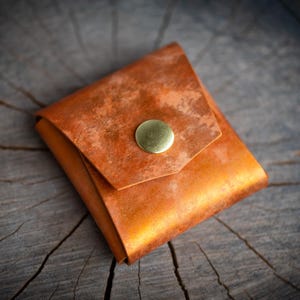 K&ouml;nnte beinhalten: Eine kleine, quadratische, braune Ledertasche mit einem Messingknopfverschluss. Die Tasche hat ein abgenutztes, verwittertes Aussehen.