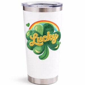 Puede incluir: Vaso de viaje blanco con base y tapa de acero inoxidable. El vaso presenta un trébol verde con la palabra "Lucky" en letras doradas, un arcoíris y un corazón verde.