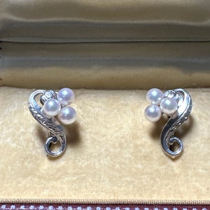 Op de afbeelding: Een paar zilveren oorbellen met een bloemmotief en drie witte parels aan elke oorbel. De oorbellen zitten in een doosje met het Mikimoto Pearl logo.