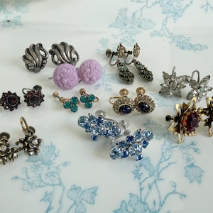 May include: A collection of ten vintage screw-back earrings featuring various designs, including silver-toned metal, lavender floral plastic, clear rhinestones, deep red glass, blue crystals, and dark metal settings with intricate floral and star motifs.