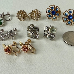 May include: A collection of vintage earrings in various styles. Includes screw-back and post-back designs. Styles include green stone drops, pearl-centered flowers, blue rhinestone flowers, clear rhinestone clusters, and rose-shaped earrings. A quarter is included for size reference.