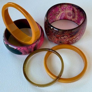 Puede incluir: Una colección de brazaletes y pulseras coloridas. La imagen presenta una mezcla de estilos, incluyendo un brazalete naranja grueso, un brazalete veteado rosa y negro, un brazalete con estampado de cachemira y dos pulseras más delgadas y translúcidas en tonos marrón y naranja.