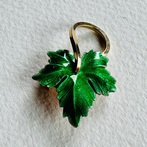 May include: A green enamel leaf pendant with a gold-tone loop for hanging. The leaf has a detailed design and a white enamel accent.
