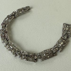 May include: Silver-toned bracelet with an Art Deco design. The bracelet features a series of rectangular and leaf-shaped links, each adorned with clear, sparkling rhinestones. The clasp is visible, and the bracelet is set against a neutral background.